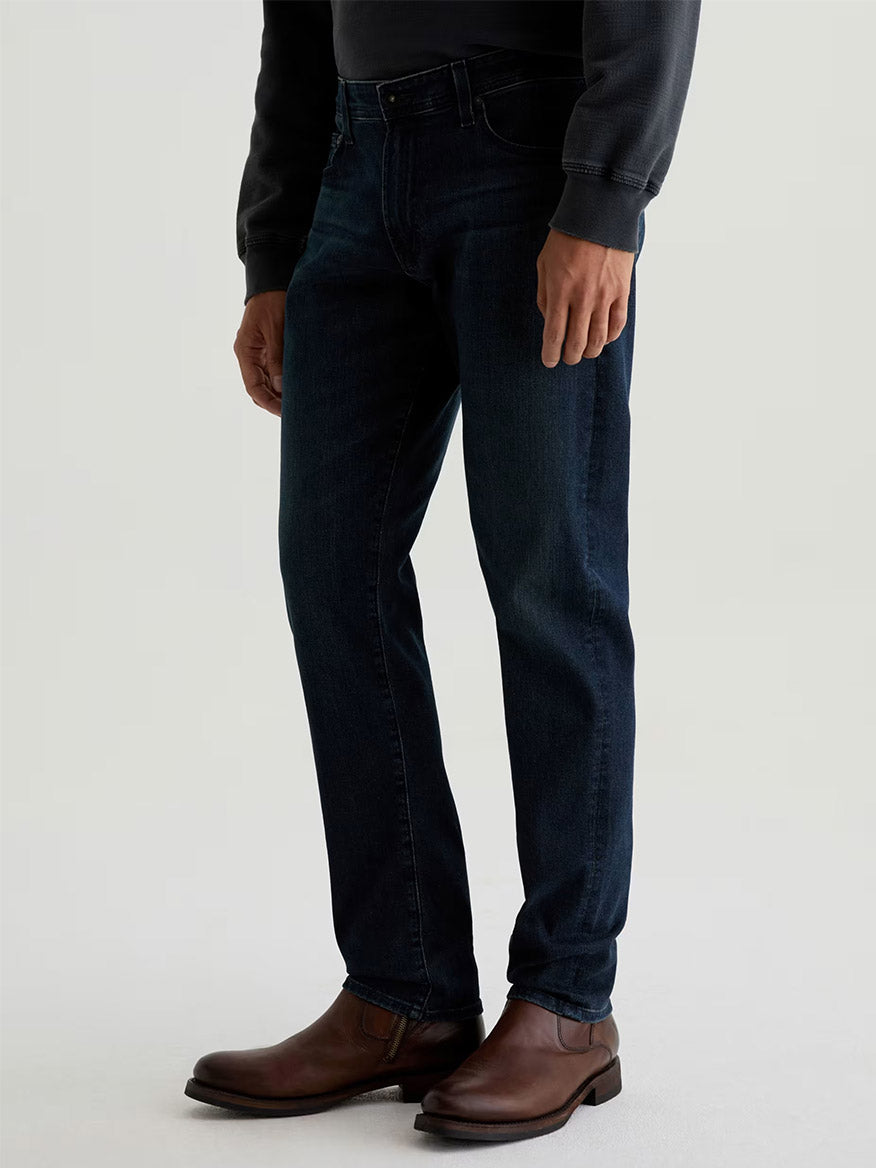 Wearing AG Jeans Tellis in Bishop, a dark indigo slim fit, paired with a long-sleeve dark shirt and brown leather boots, a person stands against a plain light background.