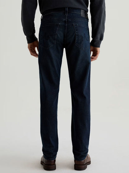 A man, seen from the back against a plain light background, wears AG Jeans Tellis in Bishop—a slim fit in dark indigo—paired with a dark long-sleeve shirt and brown shoes.