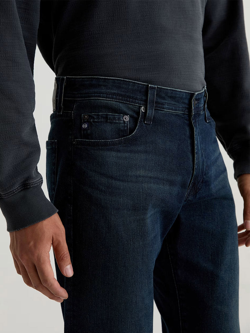 A person is wearing a dark sweatshirt and AG Jeans Tellis in Bishop slim indigo jeans, with one hand relaxed at their side.