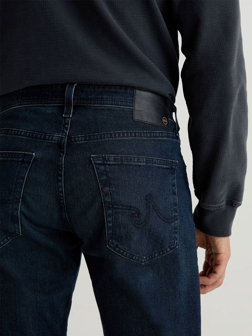 A person is seen from the back wearing the AG Jeans Tellis in Bishop, a dark indigo slim style with visible stitching and a leather patch on the waistband, paired with a dark long-sleeve shirt.