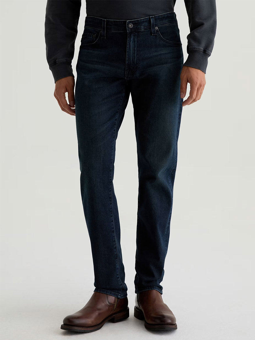 Someone wearing AG Jeans Tellis in Bishop—a dark indigo wash slim jean—paired with a dark long-sleeve shirt and brown leather boots stands against a plain light background.