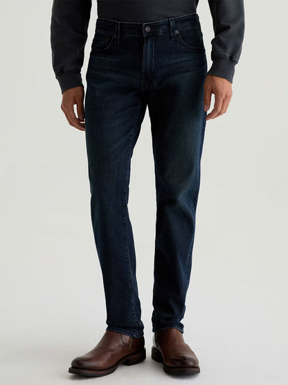 Someone wearing AG Jeans Tellis in Bishop—a dark indigo wash slim jean—paired with a dark long-sleeve shirt and brown leather boots stands against a plain light background.