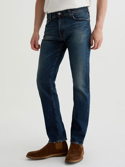 A person wearing AG Jeans Tellis in 9 Years Alton, paired with a white shirt and brown suede boots, stands against a plain light background.