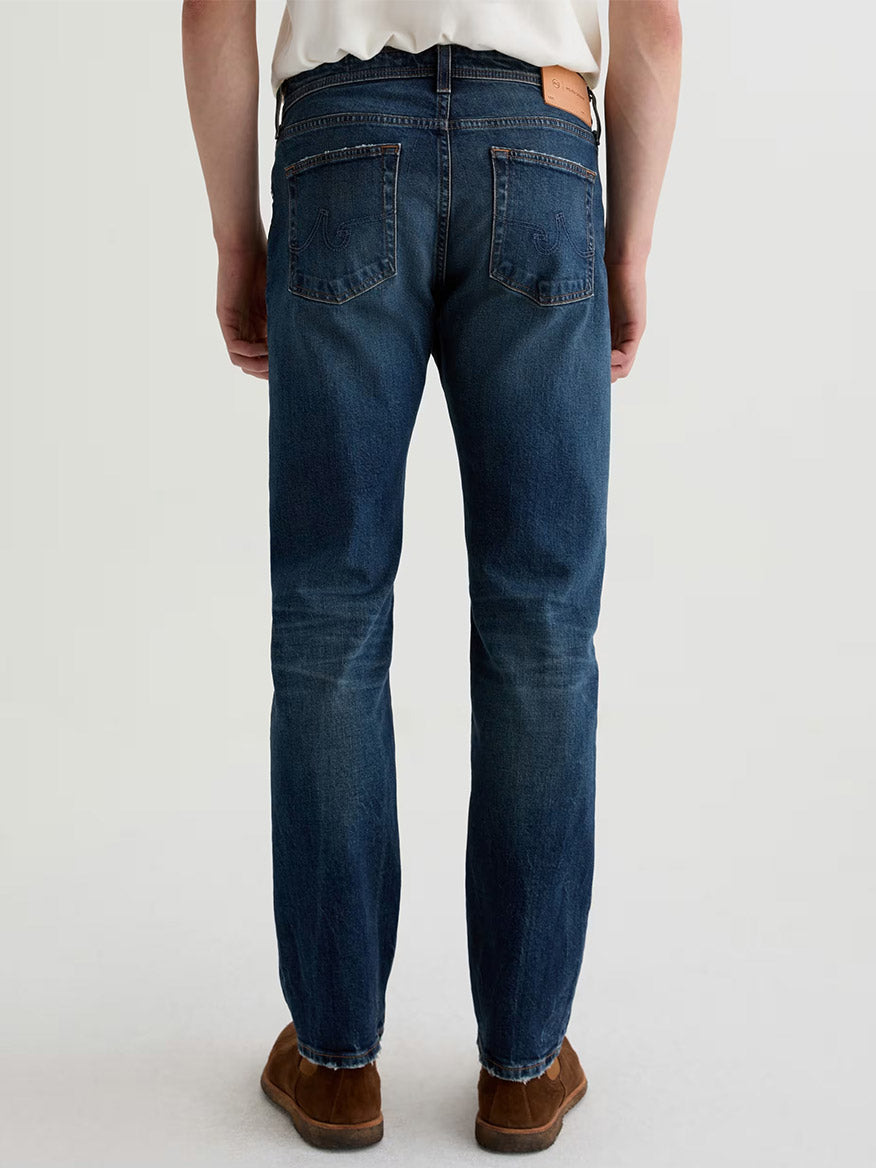 Someone wearing AG Jeans Tellis in 9 Years Alton, paired with a white shirt and brown shoes, stands facing away from the camera against a plain background.