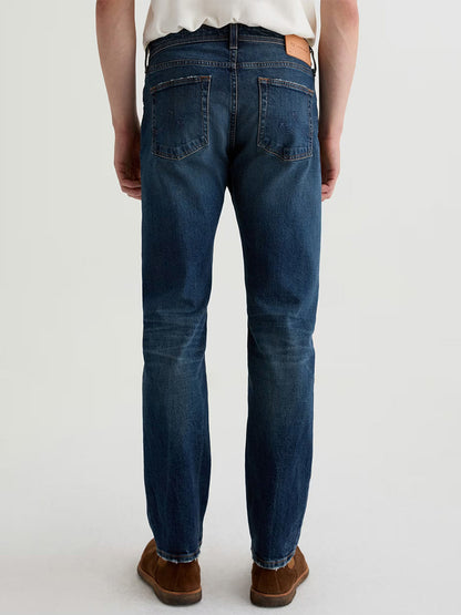 Someone wearing AG Jeans Tellis in 9 Years Alton, paired with a white shirt and brown shoes, stands facing away from the camera against a plain background.