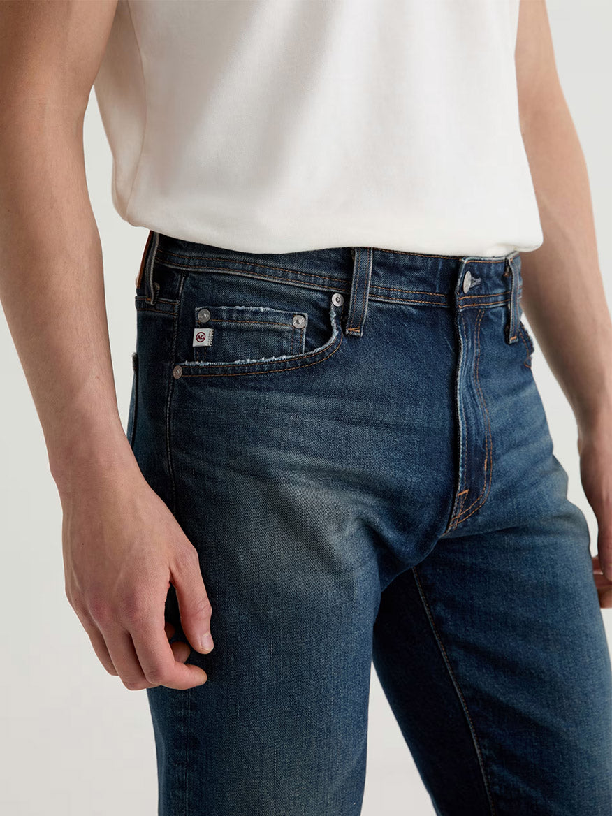Model wears the AG Jeans Tellis in 9 Years Alton, a blue premium denim slim-fit jean, paired with a white shirt. Shown from shoulders to thighs, with one arm relaxed at their side.
