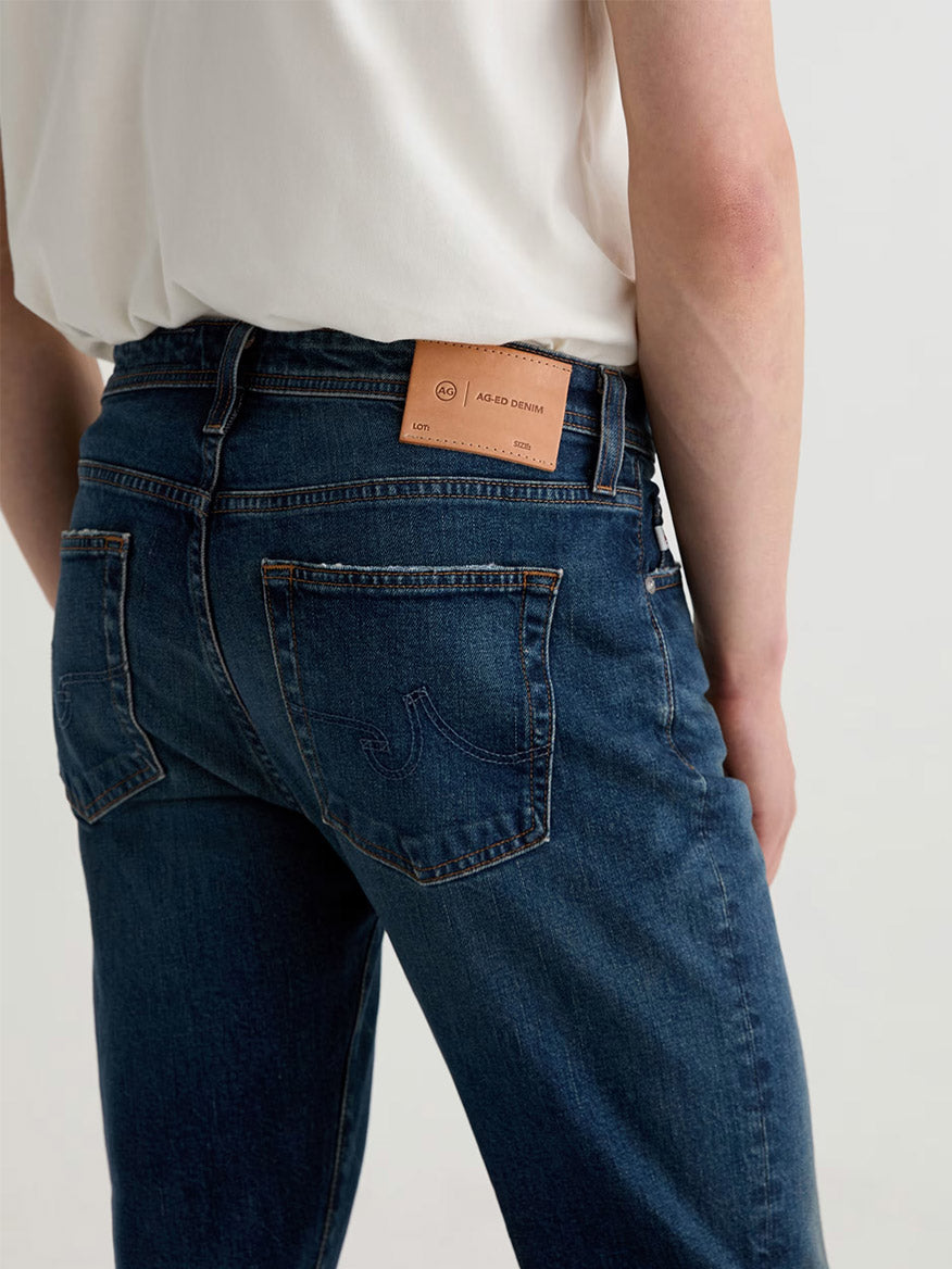 A person wears AG Jeans Tellis in 9 Years Alton—modern slim blue jeans with a white shirt, shown from the back; a tan leather brand patch above the right pocket showcases premium denim craftsmanship.