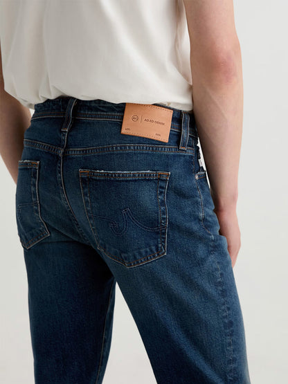A person wears AG Jeans Tellis in 9 Years Alton—modern slim blue jeans with a white shirt, shown from the back; a tan leather brand patch above the right pocket showcases premium denim craftsmanship.