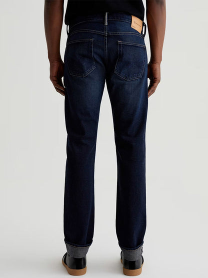 A person faces away, wearing AG Jeans Tellis in 9 Years Robust—slim-fit selvage denim—paired with a black shirt and black shoes with brown soles, set against a plain light background.