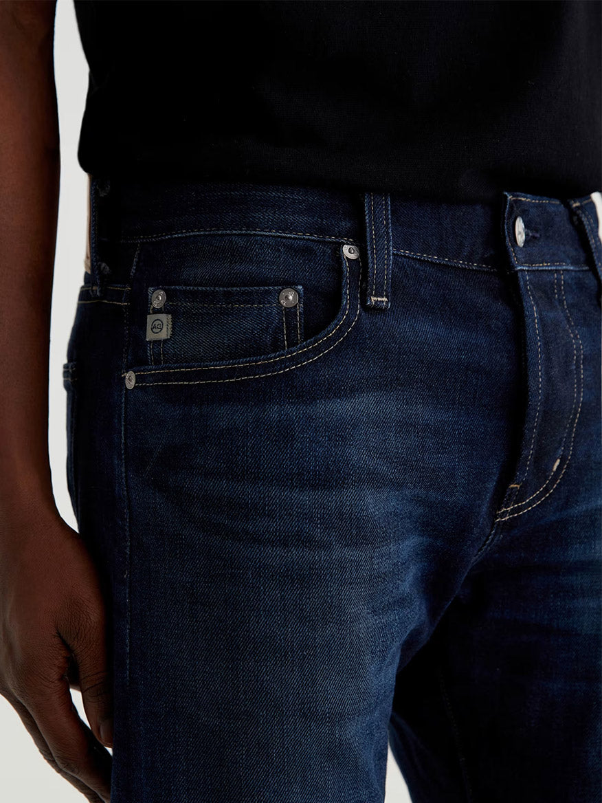 A person wearing AG Jeans Tellis in 9 Years Robust, dark blue slim-fit jeans made from organic cotton, paired with a black shirt. Shown from waist to mid-thigh with a hand resting by the side.