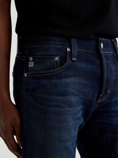 A person wearing AG Jeans Tellis in 9 Years Robust, dark blue slim-fit jeans made from organic cotton, paired with a black shirt. Shown from waist to mid-thigh with a hand resting by the side.