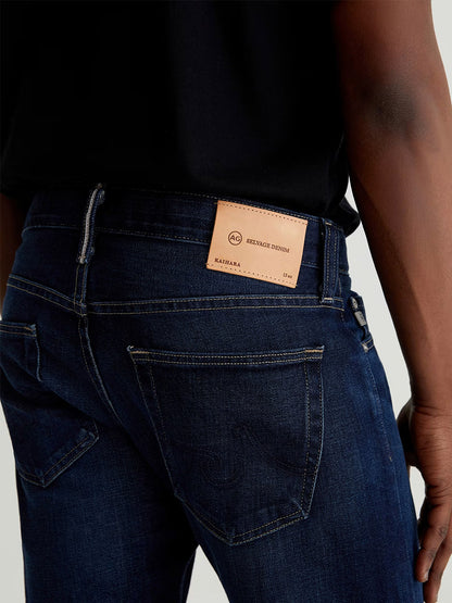 A person wearing AG Jeans Tellis in 9 Years Robust, slim-fit dark blue denim jeans with a leather brand label visible on the waistband above the back pocket, paired with a black shirt.