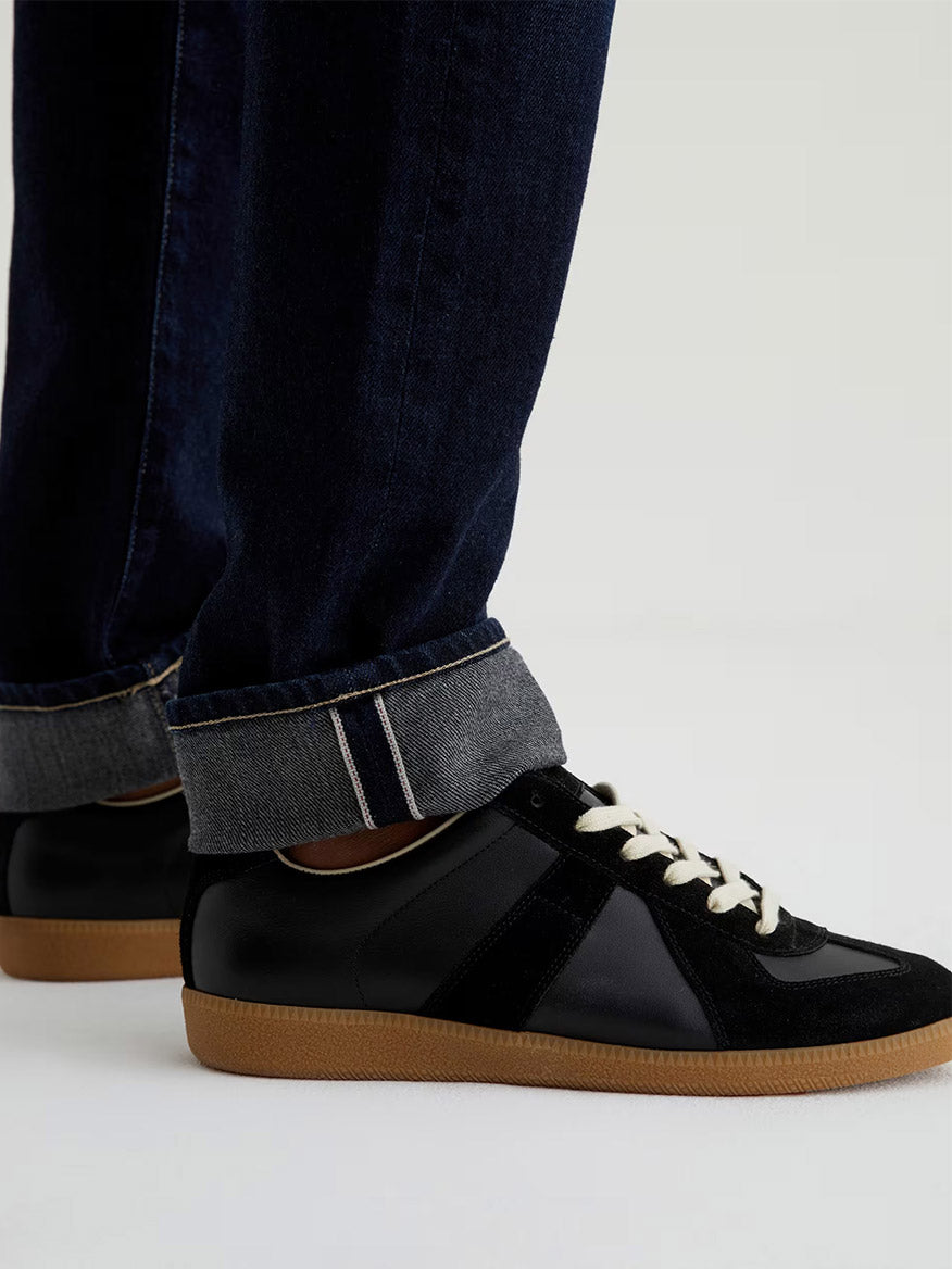 A person wearing AG Jeans Tellis in 9 Years Robust and black sneakers with white laces and gum soles, standing on a light surface.