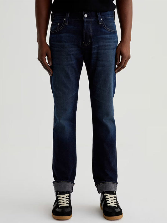 A person wearing AG Jeans Tellis in 9 Years Robust, a black shirt, and black and white sneakers stands facing forward against a plain background.