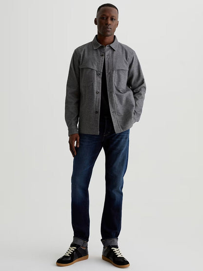 A man stands facing forward in AG Jeans Tellis in 9 Years Robust, paired with a grey button-up shirt jacket and black sneakers against a plain light background.
