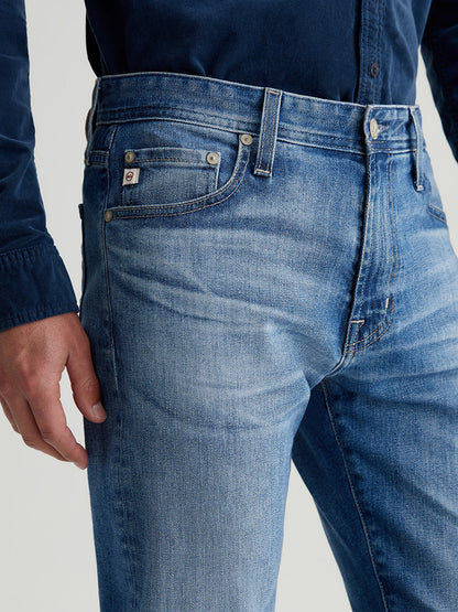 A person in a dark shirt wearing AG Jeans Tellis in 19 Years Broad, featuring visible front pocket and button details, showcasing the AG-ed™ Denim finish.