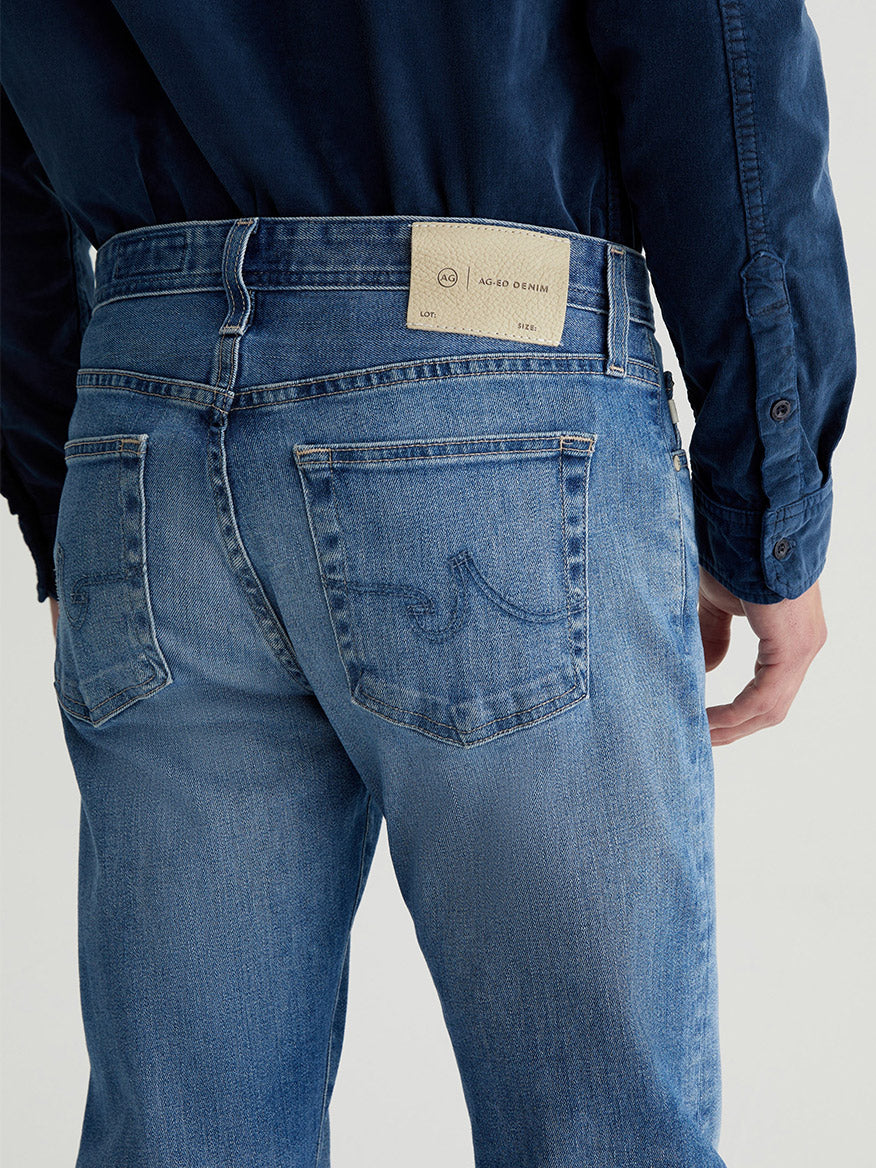 The person is wearing the AG Jeans Tellis in 19 Years Broad, featuring a label on the back pocket, paired with a dark blue shirt.