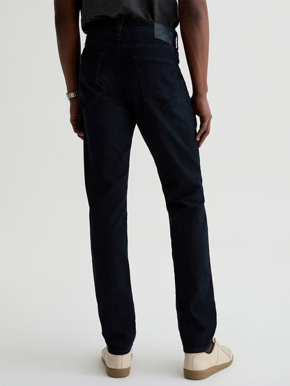A person faces away, wearing AG Jeans Tellis in Tamarack (a modern slim fit, dark blue wash), paired with a dark shirt, light sneakers, and a bracelet against a plain light background.