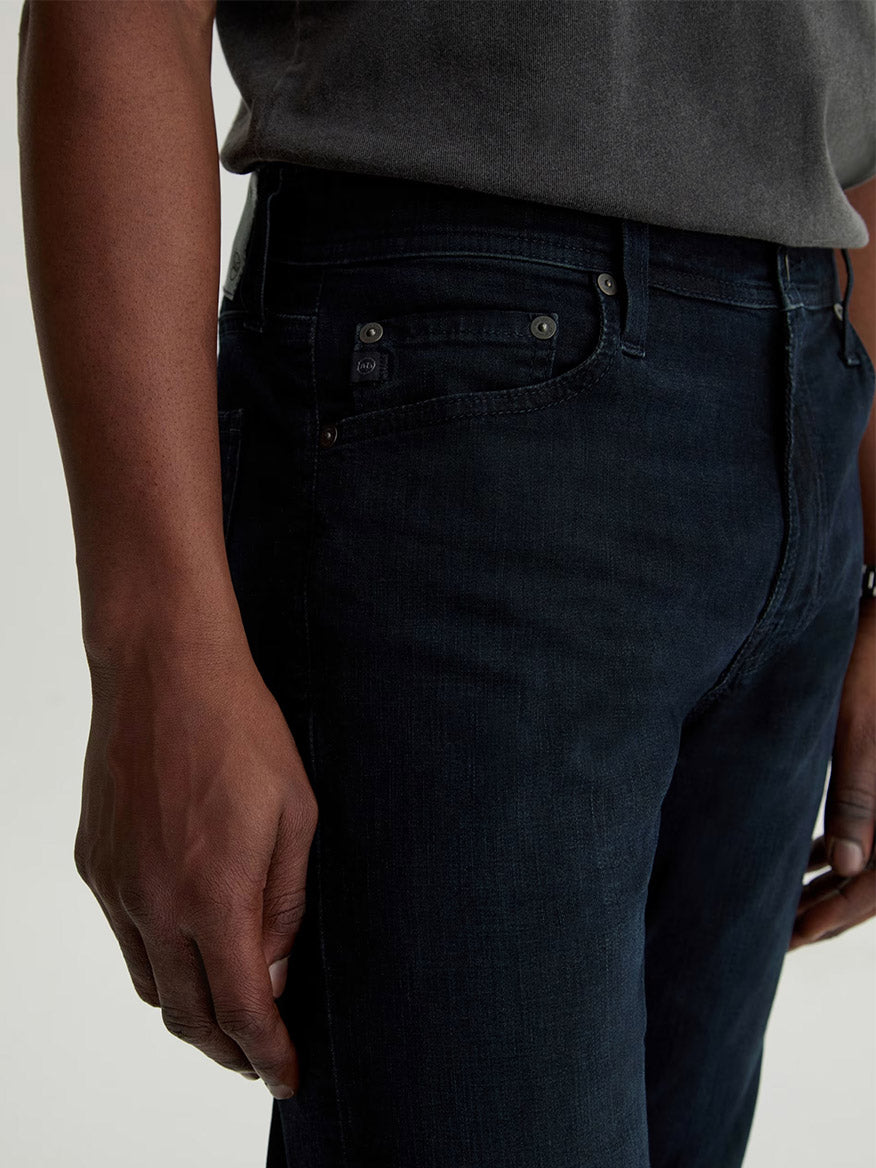 A person shown from waist to mid-thigh wears an AG Jeans Tellis in Tamarack, a modern slim jean in dark blue wash, paired with a dark gray shirt and their left hand relaxed by their side.
