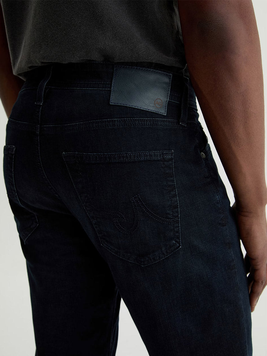 A person shown from the waist down and viewed from behind, wearing AG Jeans Tellis in Tamarack with a dark shirt.