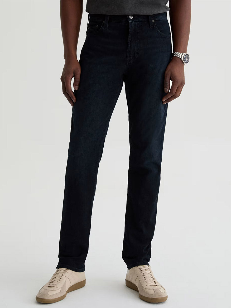 Wearing AG Jeans Tellis in Tamarack (modern slim, dark blue wash), a dark shirt, white sneakers, and a wristwatch, the person stands against a plain light background.