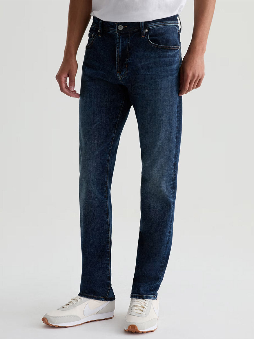 Someone wearing AG Jeans Tellis in Mastodon—a dark blue wash straight-leg denim—paired with a white t-shirt and white athletic shoes, standing against a plain light background.