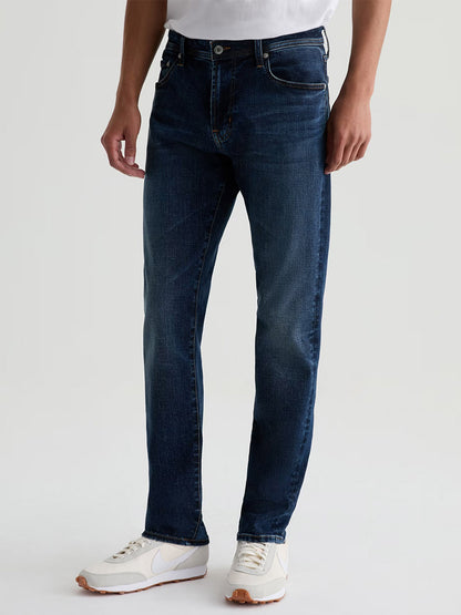 Someone wearing AG Jeans Tellis in Mastodon—a dark blue wash straight-leg denim—paired with a white t-shirt and white athletic shoes, standing against a plain light background.