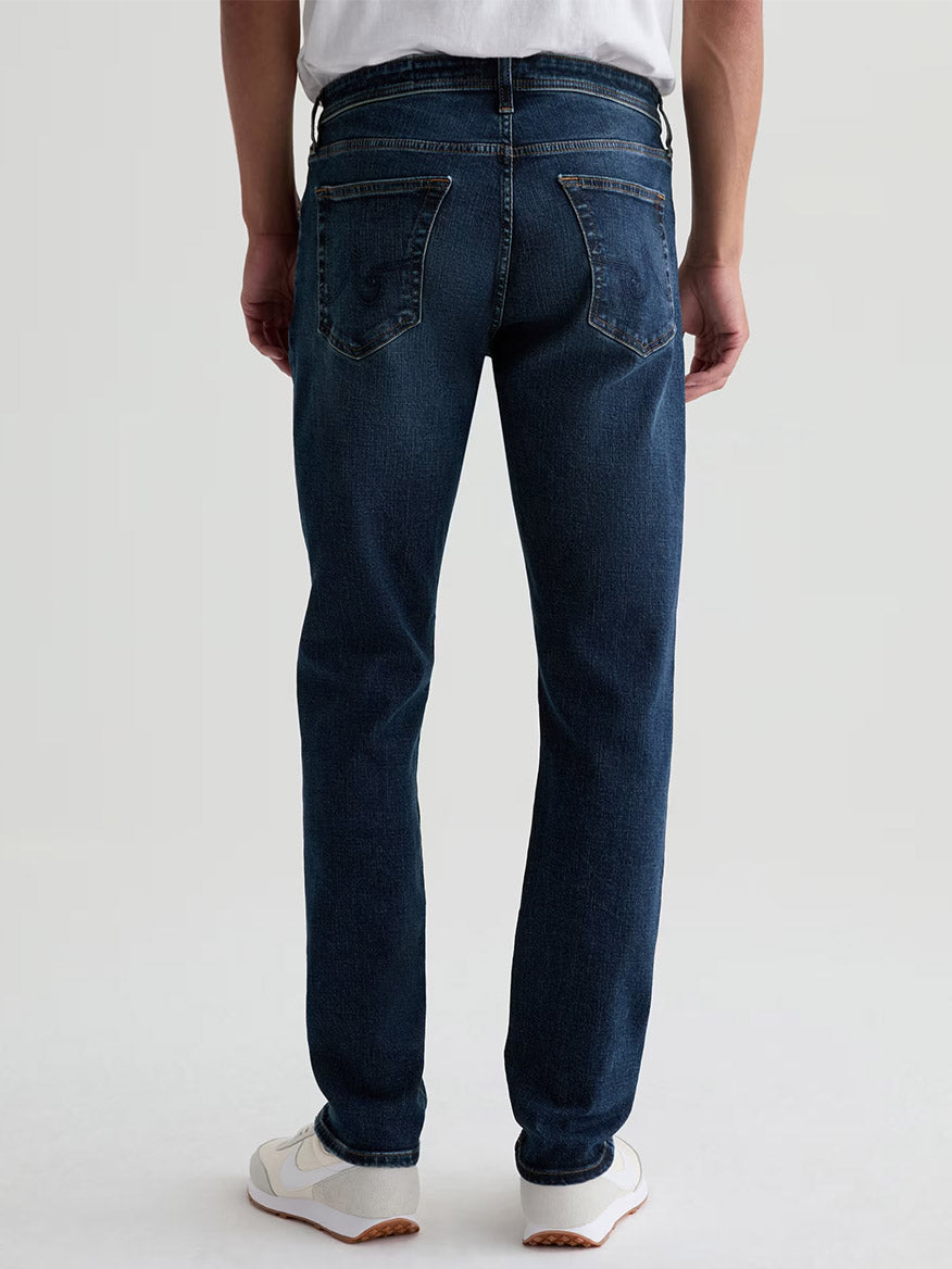 A person wearing a white shirt, AG Jeans Tellis in Mastodon (a dark blue wash modern slim jean), and white sneakers stands facing away from the camera against a plain background.