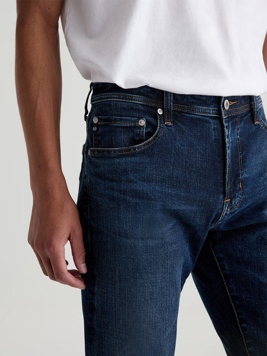 A person wears a white T-shirt and AG Jeans Tellis in Mastodon—a modern slim-fit jean in dark blue wash—shown from mid-torso to upper thigh, with one arm relaxed at their side.