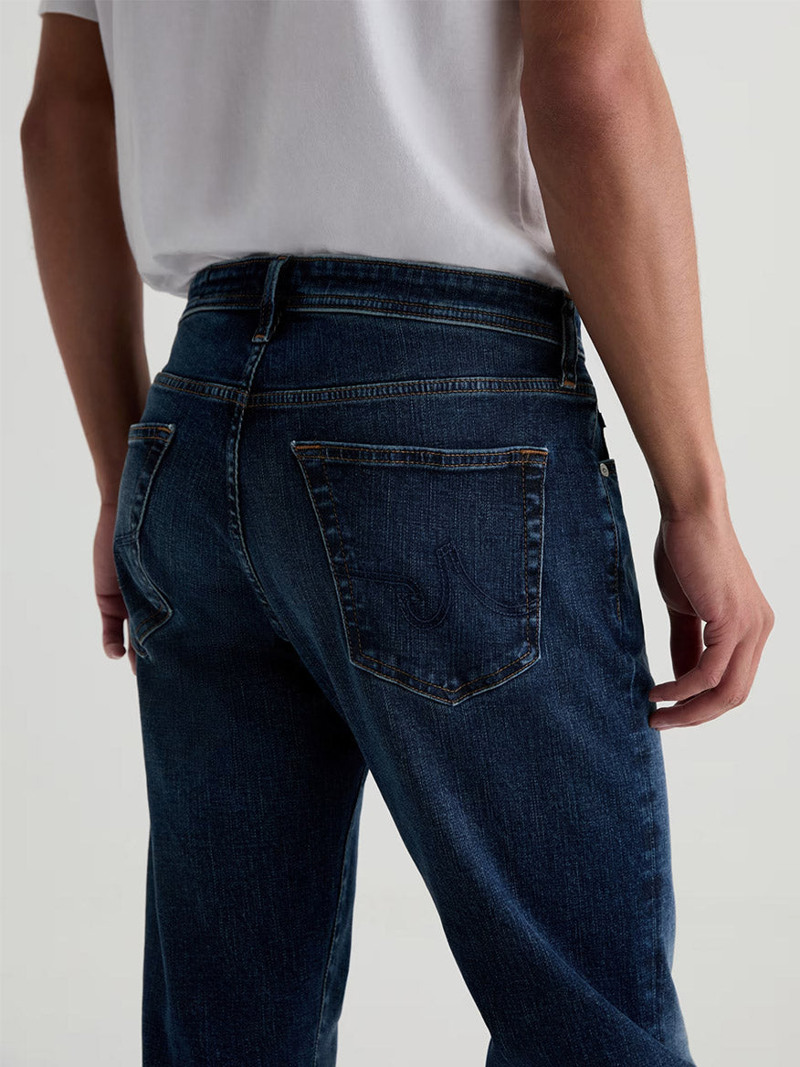 Model wearing AG Jeans Tellis in Mastodon, a modern slim dark blue wash jean, paired with a white t-shirt; shown from the back at waist height with one hand resting by their side.