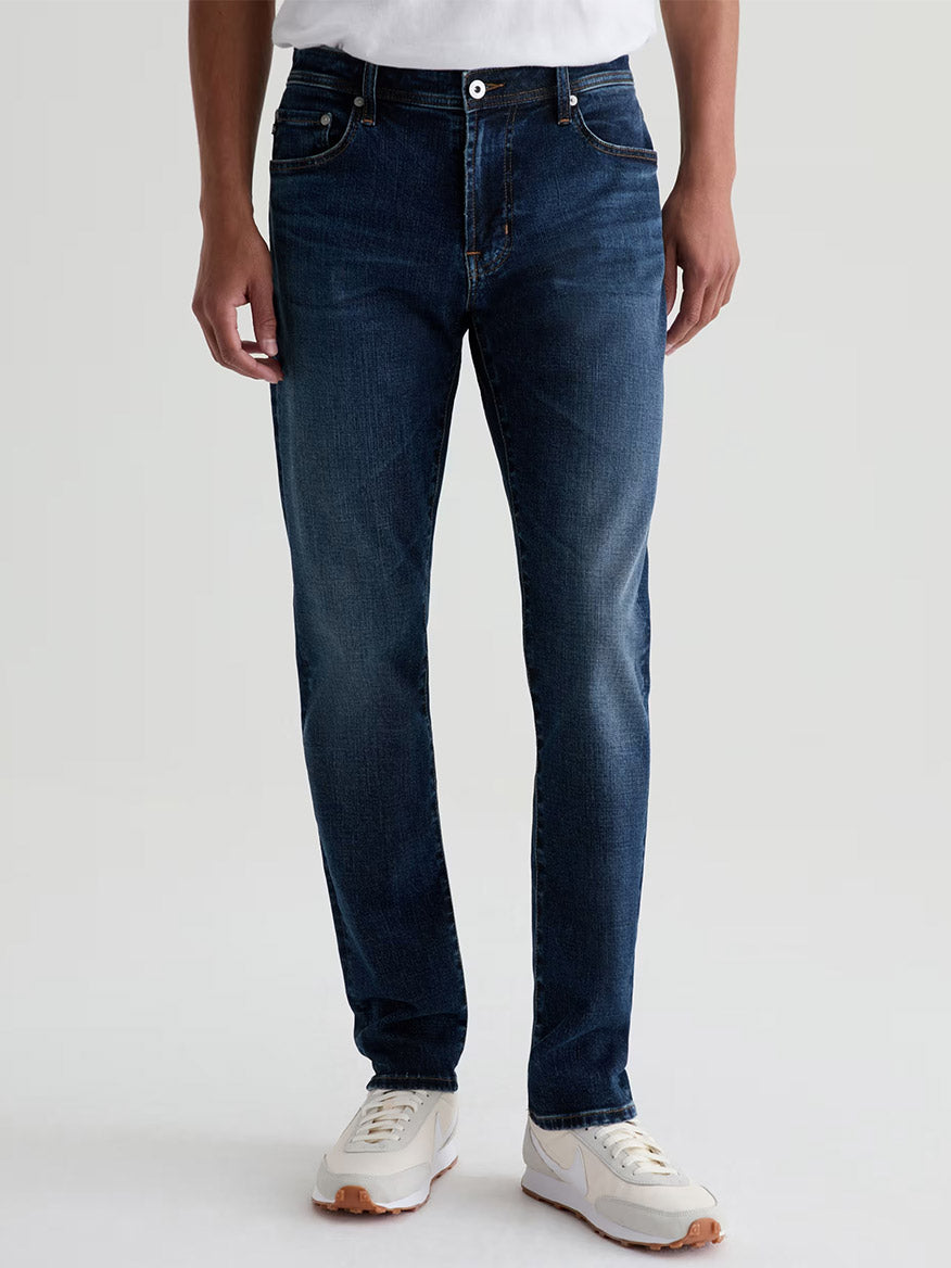 Someone wearing AG Jeans Tellis in Mastodon, a modern slim-fit jean in dark blue, paired with a white t-shirt and white sneakers, stands against a plain light background.