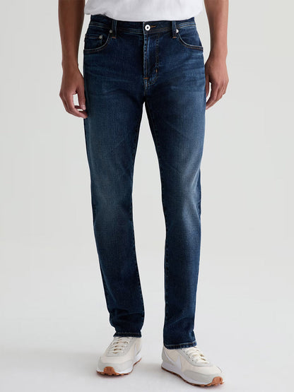 Someone wearing AG Jeans Tellis in Mastodon, a modern slim-fit jean in dark blue, paired with a white t-shirt and white sneakers, stands against a plain light background.