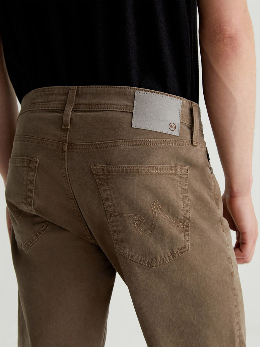 A person is wearing the AG Jeans Tellis Sueded Modern Slim in Sulfur Faded Ashwood, along with a black shirt, viewed from the back. These men's fashion jeans feature a stitched pocket design and a leather patch on the waistband, crafted from sueded stretch sateen for added comfort and style.