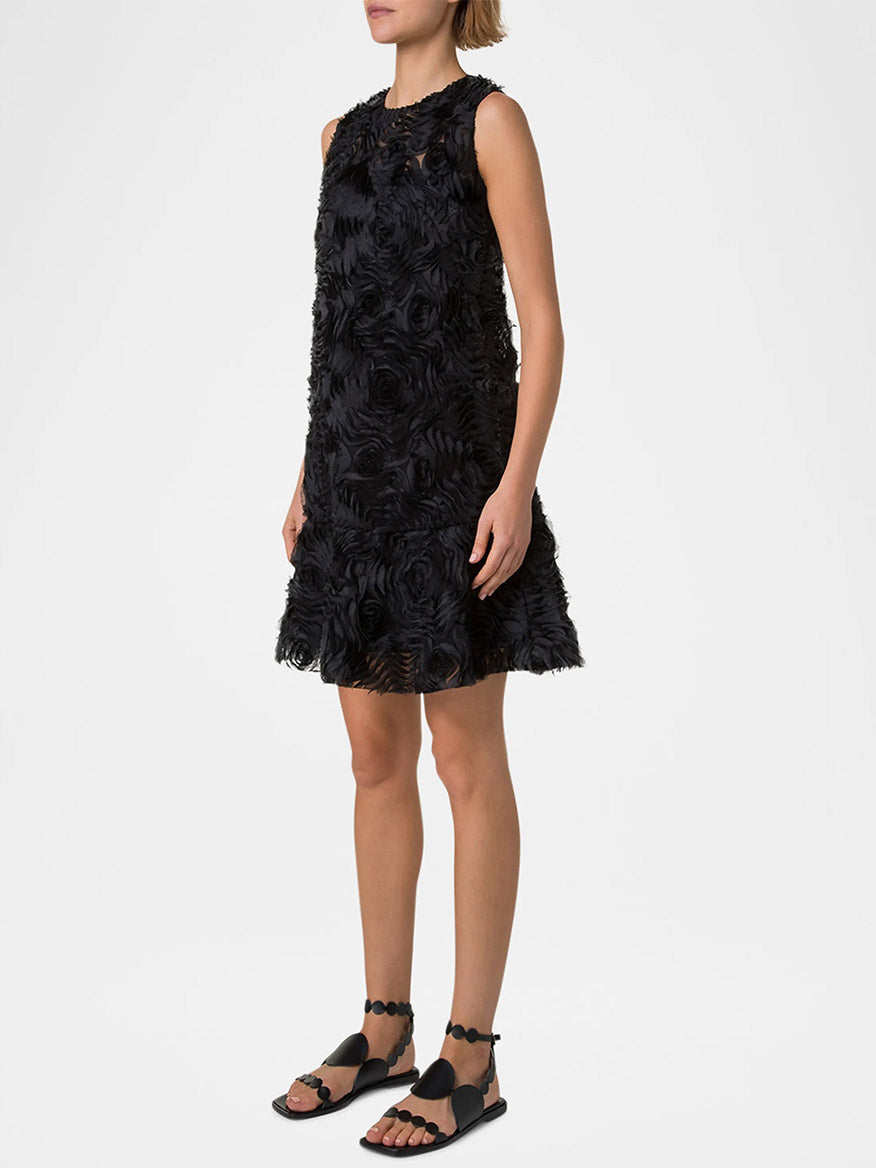 A person is wearing the Akris Punto Sleeveless 3D Carnation Knee-Length Dress in black, beautifully adorned with a textured floral design, paired perfectly with black sandals.