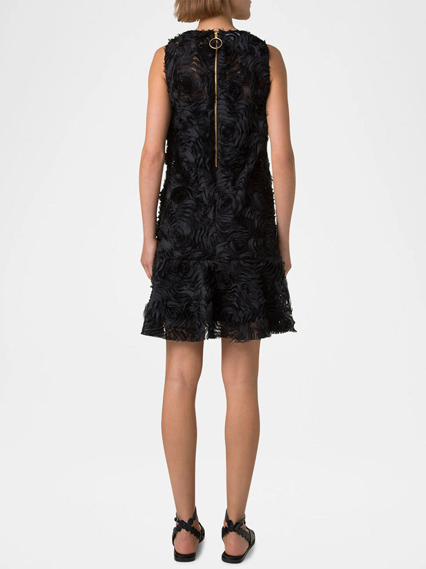 A person faces away, wearing the Akris Punto Sleeveless 3D Carnation Knee Length Dress in black, featuring textured floral patterns and a gold zipper in a chic shift silhouette, paired with black sandals.