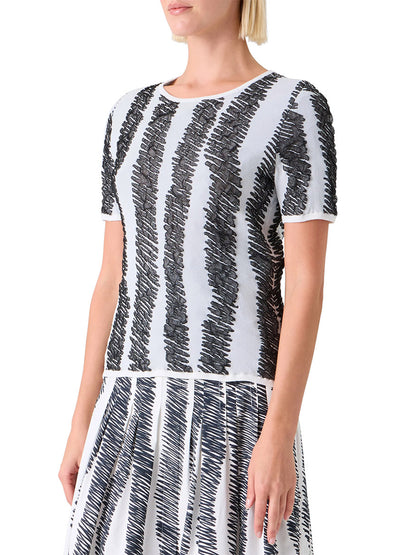 A woman wears the Akris Punto 3D Scribble Jacquard Knit Sweater in cream/black, featuring black irregular vertical stripes on a white background and crafted with stretch fibers for comfort.