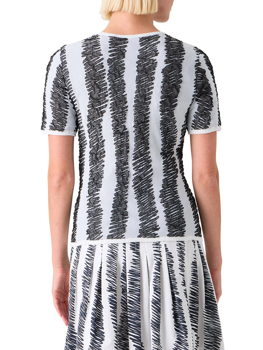 A person with short blonde hair, seen from the back, wears the Akris Punto 3D Scribble Jacquard Knit Sweater in Cream/Black, featuring black scribble-like vertical stripes on a light background.