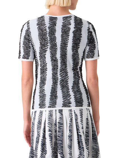 A person with short blonde hair, seen from the back, wears the Akris Punto 3D Scribble Jacquard Knit Sweater in Cream/Black, featuring black scribble-like vertical stripes on a light background.