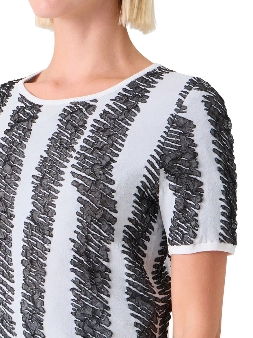 A person wearing the Akris Punto 3D Scribble Jacquard Knit Sweater in Cream/Black, featuring short sleeves and raised black textured stripes. The image shows their upper torso, neck, and part of their face.