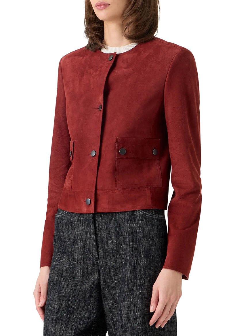A woman wears the Akris Punto Boxy Suede Jacket in Pomegranate, a collarless, button-front goatskin suede piece with utility pockets, paired with dark textured pants.