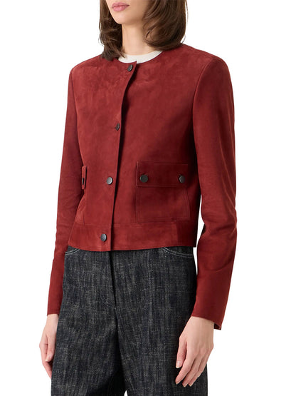 A woman wears the Akris Punto Boxy Suede Jacket in Pomegranate, a collarless, button-front goatskin suede piece with utility pockets, paired with dark textured pants.