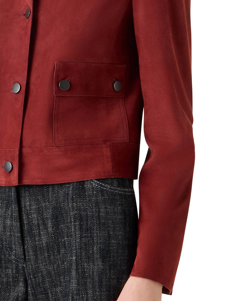 A person wearing the Akris Punto Boxy Suede Jacket in Pomegranate, featuring utility-inspired pockets, paired with dark textured pants, shown from shoulder to mid-thigh.