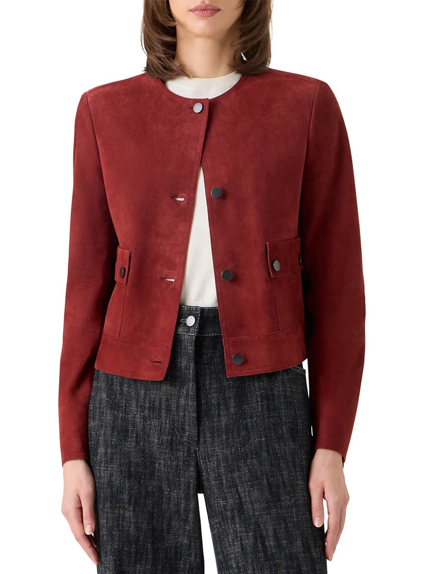 A person wears the Akris Punto Boxy Suede Jacket in Pomegranate, featuring utility-inspired pockets and a jewel neckline, layered over a white top and dark textured pants; photographed from the waist up.