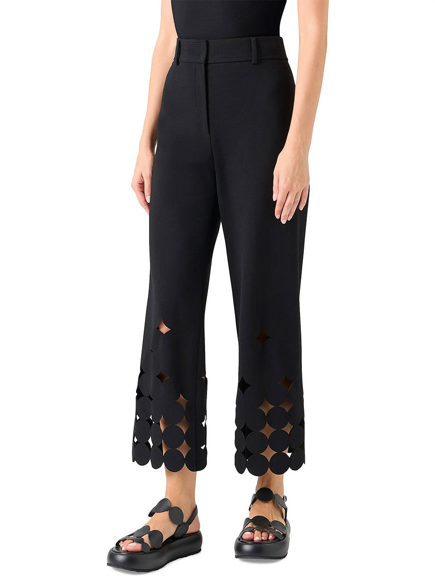 A person wearing Akris Punto Chieko Dropping Dot Cut Out Ankle Pant in black with geometric dot cutouts near the hem and black sandals, standing against a white background.