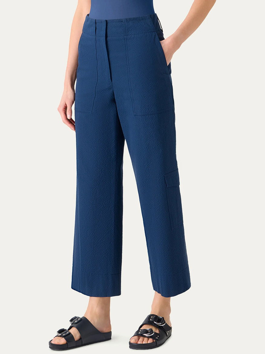 A person wears Akris Punto Cliff Seersucker Cargo Pants in Dark Blue featuring a high-waisted, wide-leg cargo style with front pockets and black slide sandals, standing against a plain light background.