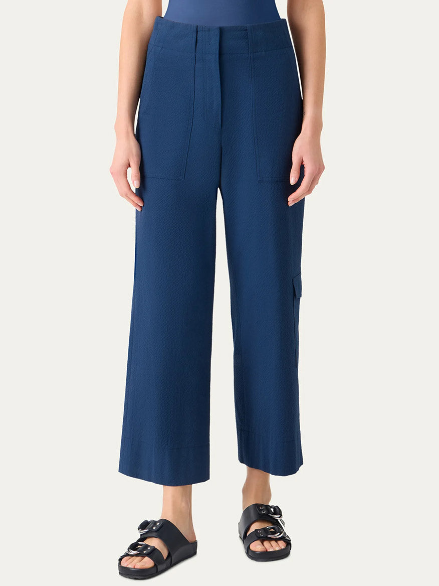 Person wears Akris Punto Cliff Seersucker Cargo Pants in Dark Blue, featuring a high-waisted, wide-leg cargo style with front pockets, paired with a tucked-in top and black slip-on sandals against a plain background.