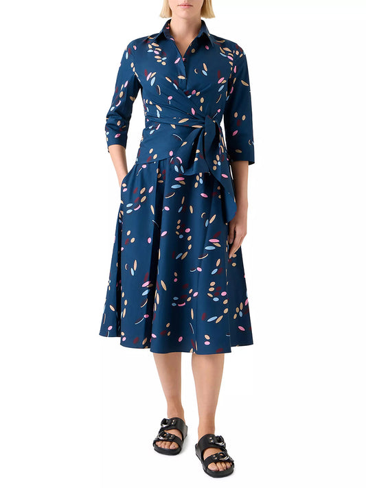 A person wearing the Akris Punto Confetti Print Wrap Dress in Ink Multi, featuring a colorful confetti pattern, three-quarter sleeves, a tied waist, and black sandals.