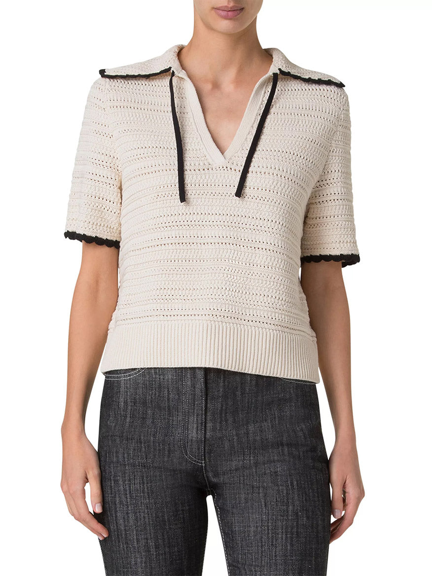 The person is wearing an Akris Punto Crochet Boxy Polo in Cashew/Black, featuring a crochet-knit design with short sleeves, a collar, and contrast scallop trim, paired with dark jeans.