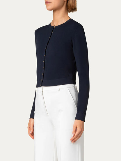 A person in an Akris Punto Cropped Cardigan with Gold Buttons in navy and white high-waisted trousers stands in profile against a plain backdrop.