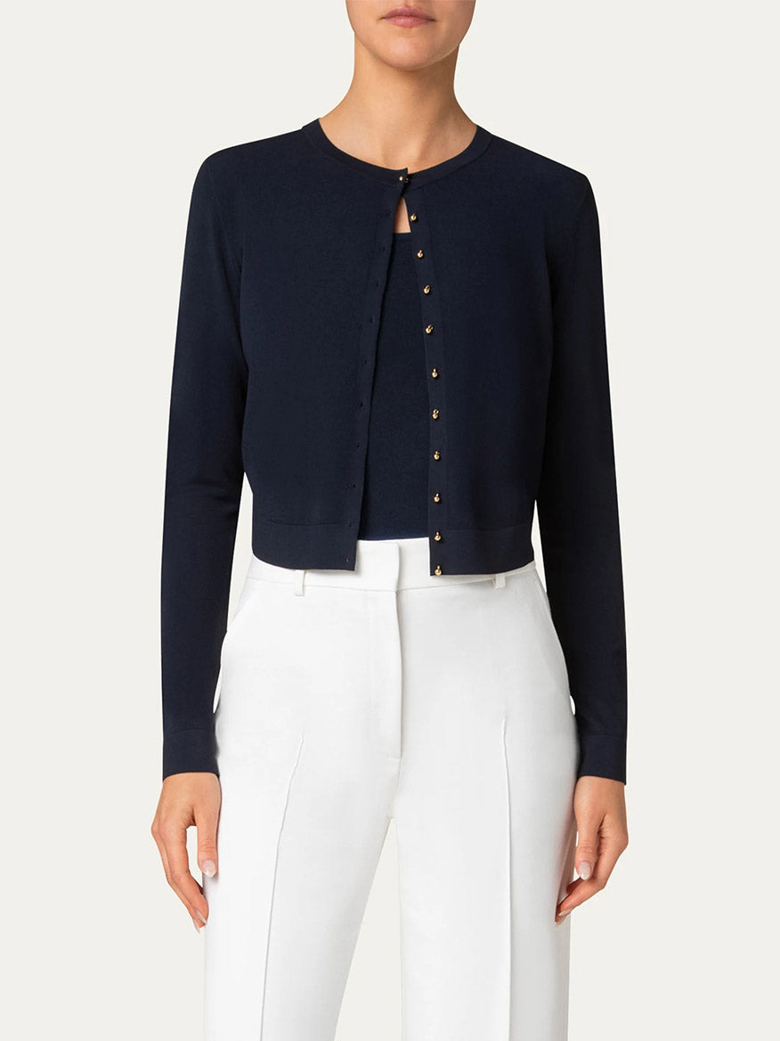 Person wearing the Akris Punto Cropped Cardigan with Gold Buttons in Navy over a white top and high-waisted white trousers, standing against a plain background.